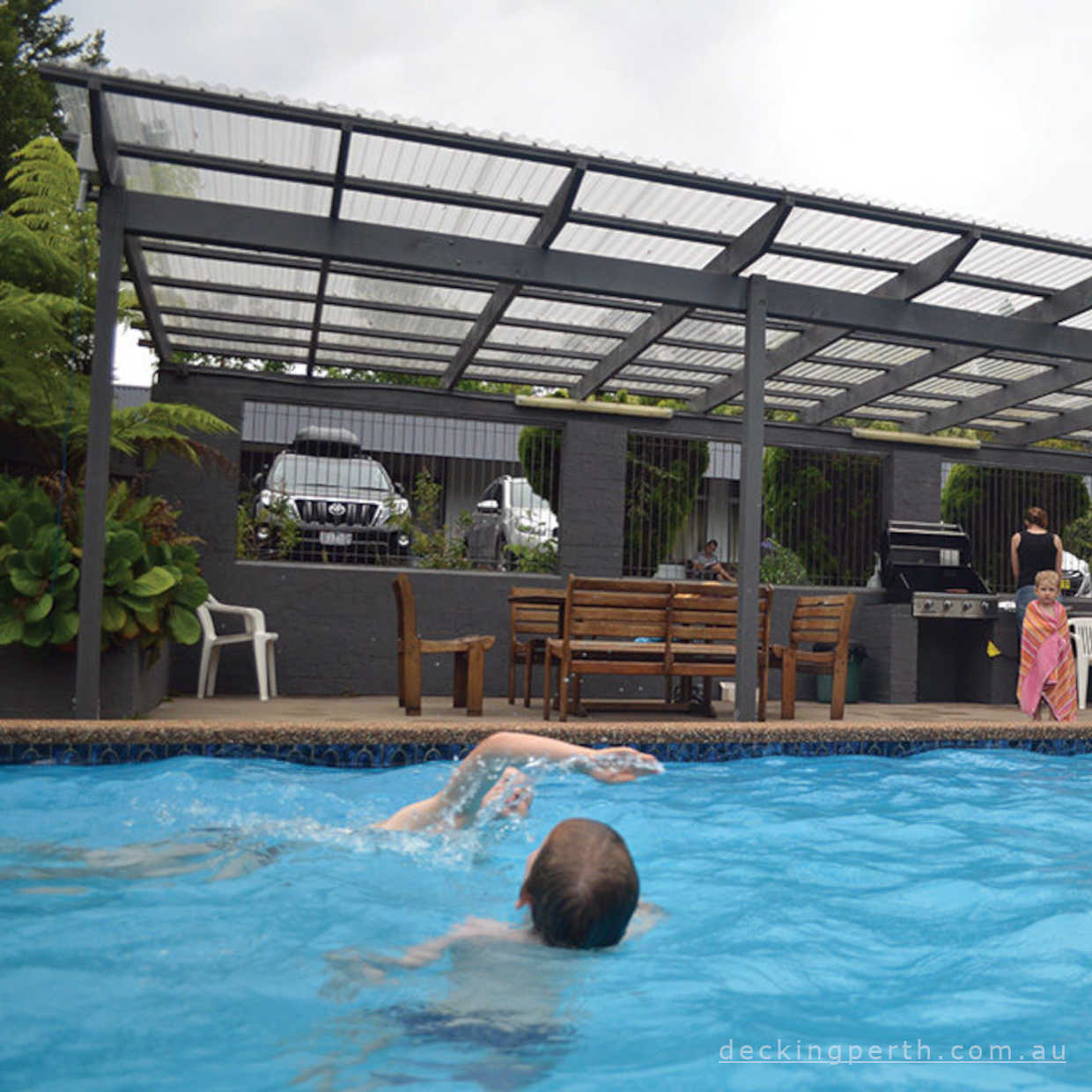 Person swimming in a pool with a pergola and outdoor furniture in the background.