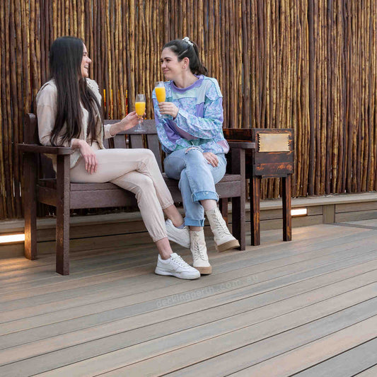 Two women sitting on a wooden bench, enjoying drinks against a rustic wall.